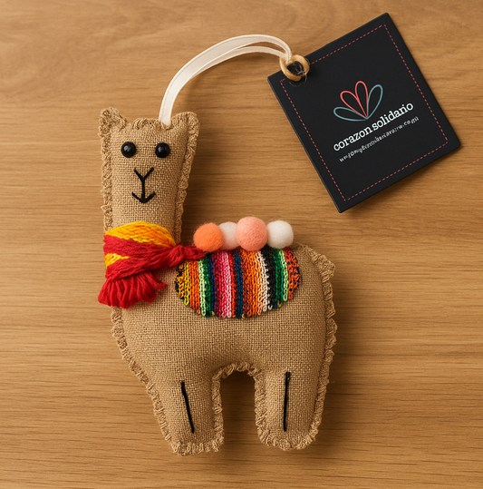 Brown, textile llama ornament with colorful scarf and pom-pom details on a wooden surface, accompanied by a 'corazones solidarios' tag.
