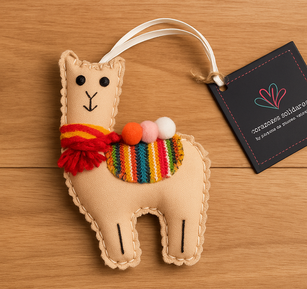 Tan llama ornament with colorful scarf and pom-poms on a wooden surface, featuring a visible brand tag.